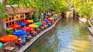 San Antonio River Walk lined with colorful umbrellas, riverside restaurants, trees, and a pedestrian bridge